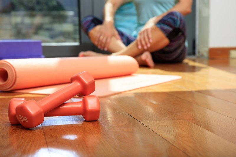 A woman in workout clothes sits on a wooden floor next to an orange yoga mat and dumbells (Photo by Rawf8 - stock.adobe.com)